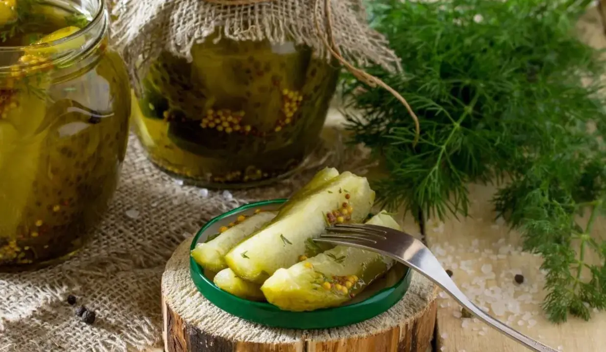 Słoiki z ogórkami w musztardzie, świeży koperek i widelec z kawałkami ogórka na talerzyku.