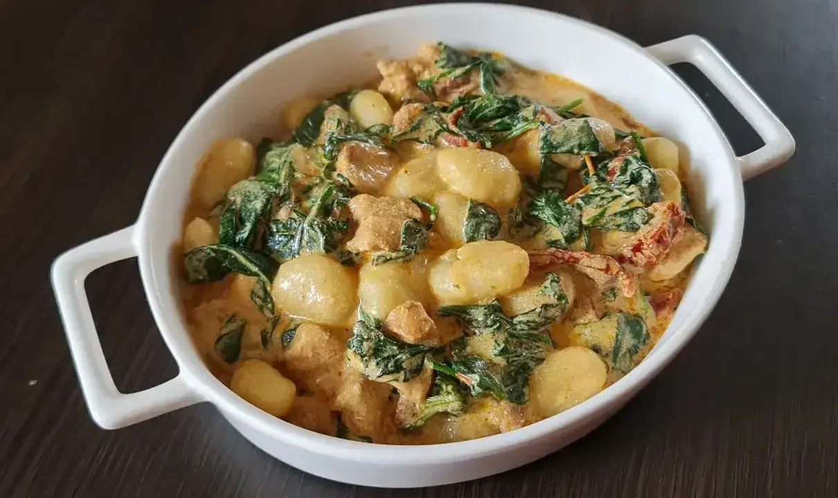Pyszne gnocchi z kurczakiem w kremowym sosie ze szpinakiem i suszonymi pomidorami. Danie podane w białej miseczce.