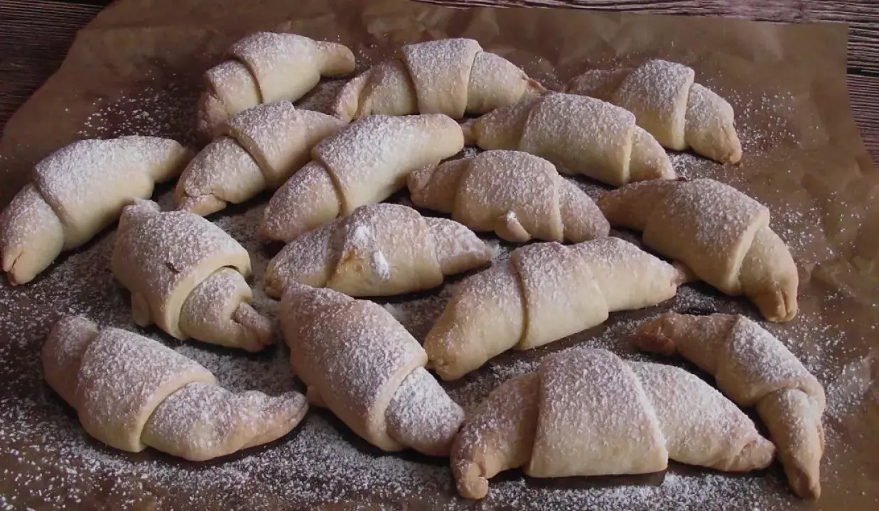 Pyszne rogaliki serowe 3 składnikowe, posypane cukrem pudrem, ułożone na papierze do pieczenia.