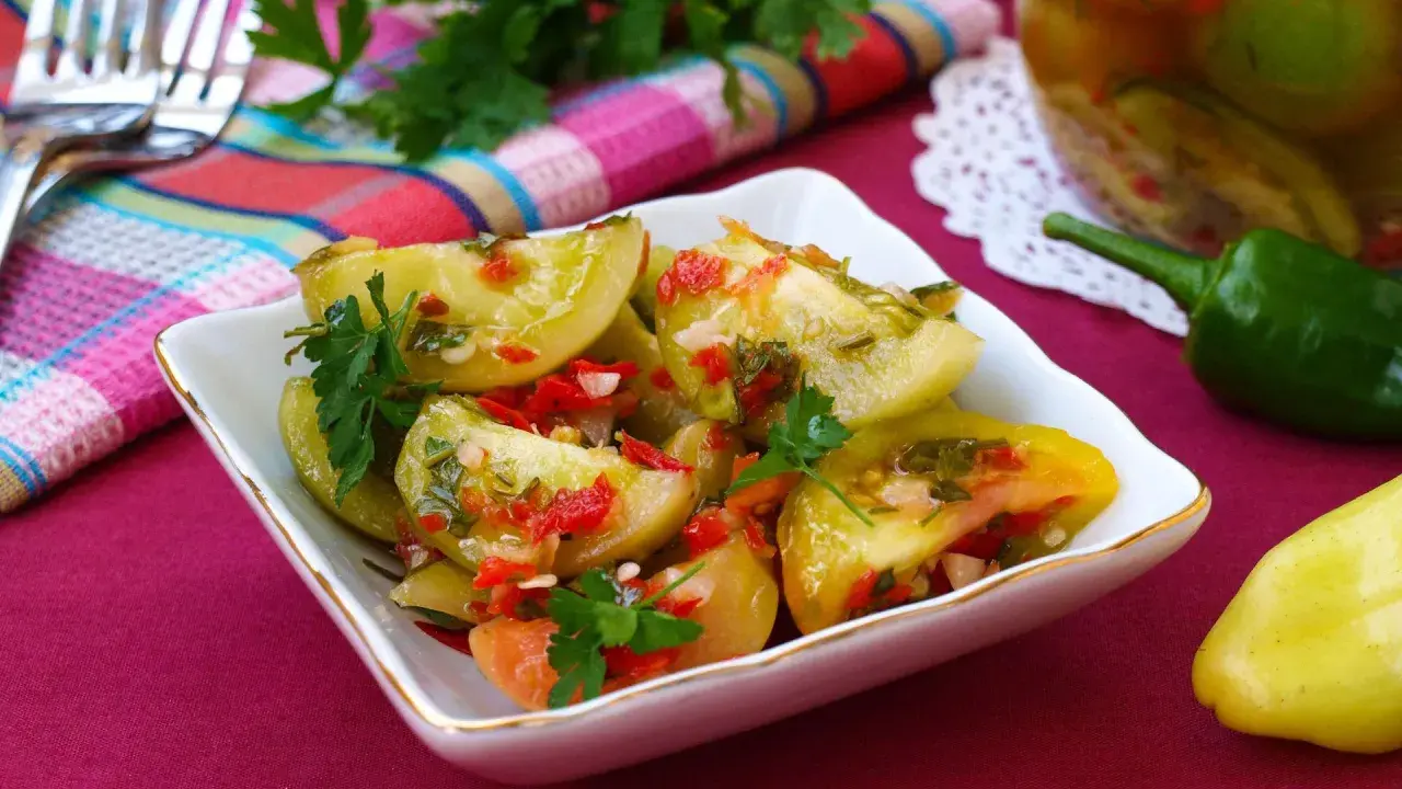 Pikantna sałatka z zielonych pomidorów z papryczką chili i natką pietruszki, podana w białej miseczce. Obok słoik z przetworami.