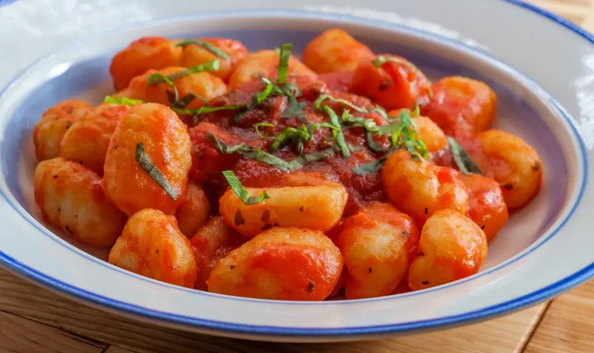 Pyszne gnocchi w sosie pomidorowym, posypane świeżą bazylią, podane w talerzu z niebieskim rantem.