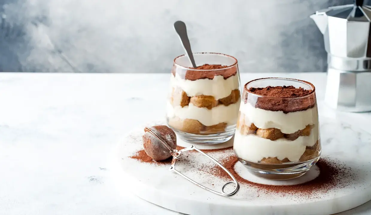Dwa pucharki z kremowym tiramisu bez jajek, przełożonym biszkoptami i posypanym kakao. Obok sitko do kakao i kawiarka.