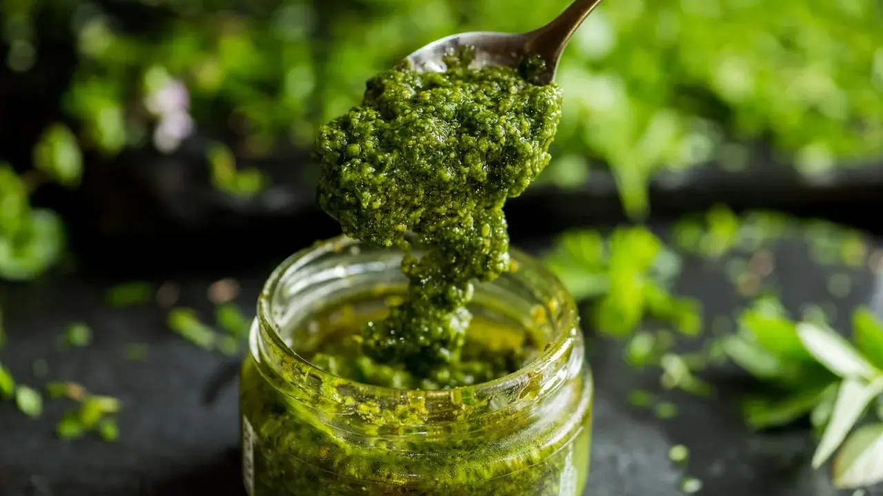 Łyżka z gęstym, zielonym pesto z pietruszki zanurza się w słoiku. W tle widać świeże zioła.