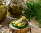 Słoiki z ogórkami w musztardzie, świeży koperek i widelec z kawałkami ogórka na talerzyku.