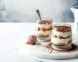 Dwa pucharki z kremowym tiramisu bez jajek, przełożonym biszkoptami i posypanym kakao. Obok sitko do kakao i kawiarka.
