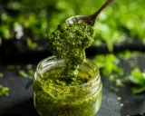 Łyżka z gęstym, zielonym pesto z pietruszki zanurza się w słoiku. W tle widać świeże zioła.