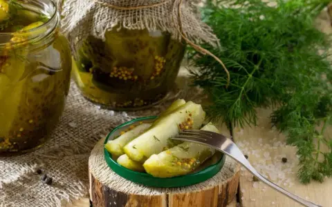 Słoiki z ogórkami w musztardzie, świeży koperek i widelec z kawałkami ogórka na talerzyku.