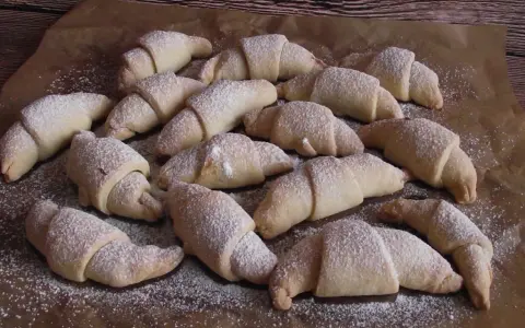 Pyszne rogaliki serowe 3 składnikowe, posypane cukrem pudrem, ułożone na papierze do pieczenia.