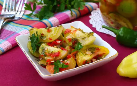 Pikantna sałatka z zielonych pomidorów z papryczką chili i natką pietruszki, podana w białej miseczce. Obok słoik z przetworami.