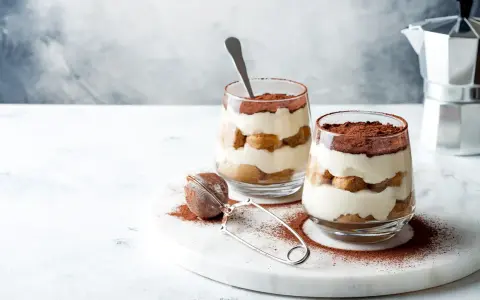Dwa pucharki z kremowym tiramisu bez jajek, przełożonym biszkoptami i posypanym kakao. Obok sitko do kakao i kawiarka.