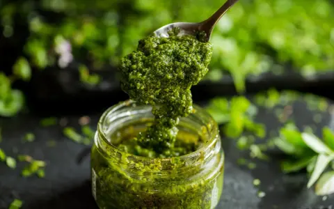 Łyżka z gęstym, zielonym pesto z pietruszki zanurza się w słoiku. W tle widać świeże zioła.