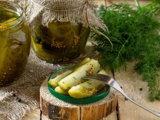 Słoiki z ogórkami w musztardzie, świeży koperek i widelec z kawałkami ogórka na talerzyku.