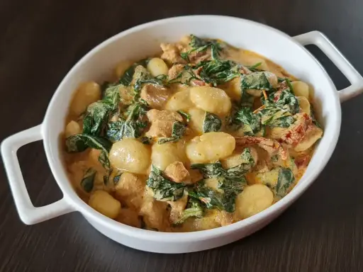 Pyszne gnocchi z kurczakiem w kremowym sosie ze szpinakiem i suszonymi pomidorami. Danie podane w białej miseczce.
