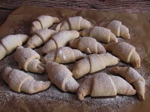 Pyszne rogaliki serowe 3 składnikowe, posypane cukrem pudrem, ułożone na papierze do pieczenia.