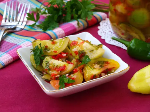 Pikantna sałatka z zielonych pomidorów z papryczką chili i natką pietruszki, podana w białej miseczce. Obok słoik z przetworami.