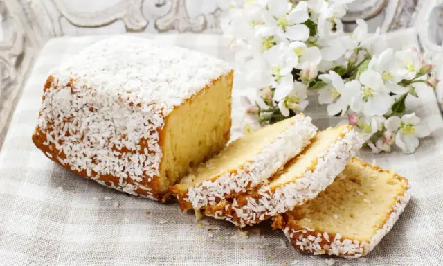 Puszyste ciasto z kokosem, posypane wiórkami, prezentuje się apetycznie obok gałązki kwitnącej jabłoni.