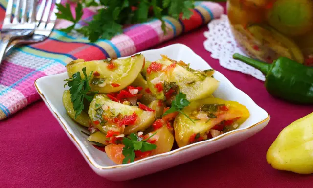 Pikantna sałatka z zielonych pomidorów z papryczką chili i natką pietruszki, podana w białej miseczce. Obok słoik z przetworami.
