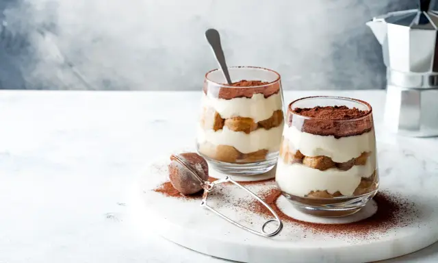 Dwa pucharki z kremowym tiramisu bez jajek, przełożonym biszkoptami i posypanym kakao. Obok sitko do kakao i kawiarka.