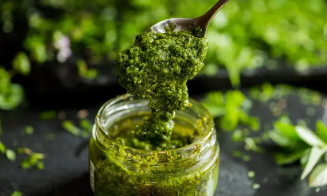 Łyżka z gęstym, zielonym pesto z pietruszki zanurza się w słoiku. W tle widać świeże zioła.