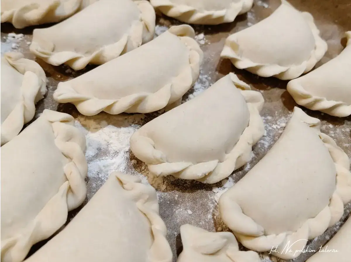 Surowe pierogi z mięsem, ułożone na blaszce, gotowe do pieczenia lub gotowania. Ich brzegi są starannie zlepione.