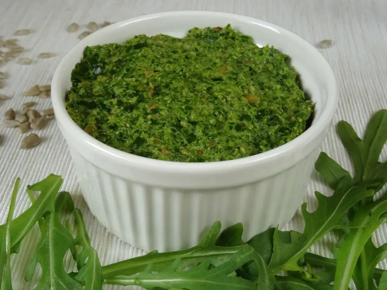 Domowe pesto z rukoli w białej miseczce, otoczone świeżymi liśćmi rukoli i nasionami słonecznika.