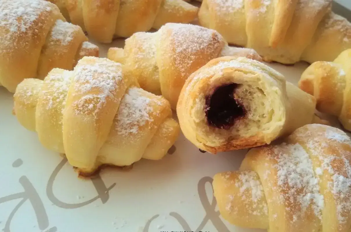 Pyszne rogaliki krucho drożdżowe, posypane cukrem pudrem. Jeden rogalik przekrojony, ukazujący ciemne, owocowe nadzienie.