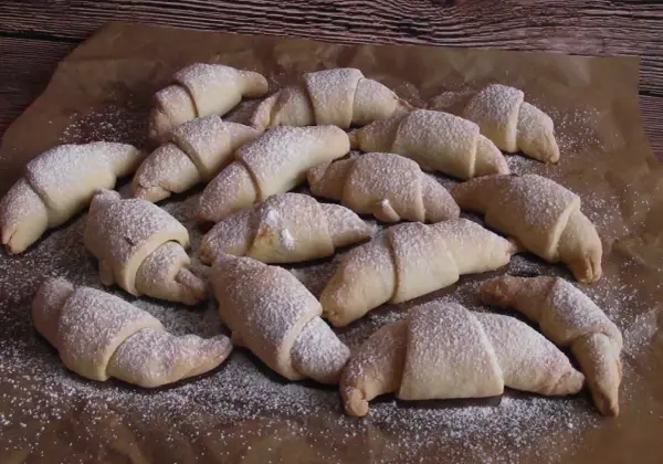 Pyszne rogaliki serowe 3 składnikowe, posypane cukrem pudrem, ułożone na papierze do pieczenia.