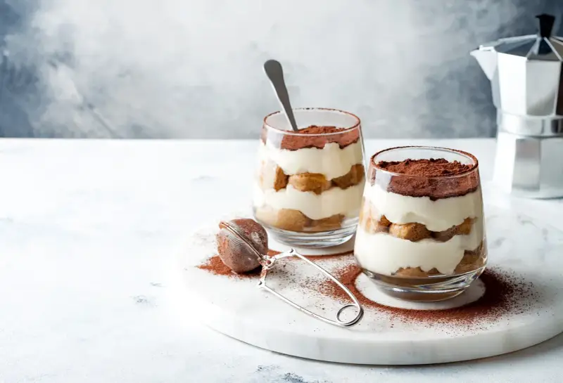 Dwa pucharki z kremowym tiramisu bez jajek, przełożonym biszkoptami i posypanym kakao. Obok sitko do kakao i kawiarka.